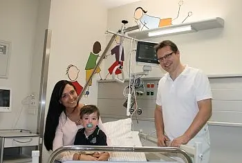 In einem Krankenhauszimmer sitzt ein kleiner Junge mit Schnuller in einem Kinderbett. Neben ihm steht ein lächelnder Arzt in weißer Kleidung, daneben eine Frau, vermutlich die Mutter. Medizinische Geräte am Bett deuten auf eine kinderfreundliche Station.