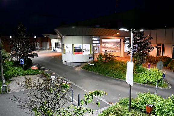 Nachtszene: Hell erleuchteter Eingangsbereich eines Krankenhauses mit runder Vorfahrt und breiter Schiebetür. Leere Zufahrt, wenige Wegweiser und Straßenlampen, umgeben von Sträuchern.