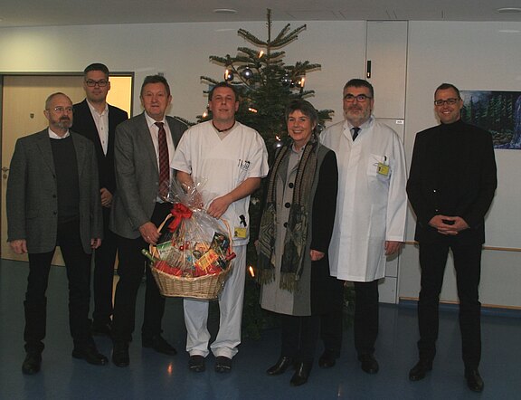 Gruppenfoto in einem Krankenhaus: Mehrere Mitarbeitende stehen vor einem Weihnachtsbaum. In der Mitte hält eine Pflegekraft einen großen Geschenkkorb mit roter Schleife. Daneben stehen Personen in Anzügen und zwei in weißen Kitteln. Festliche Stimmung, Übergabe eines Präsents.