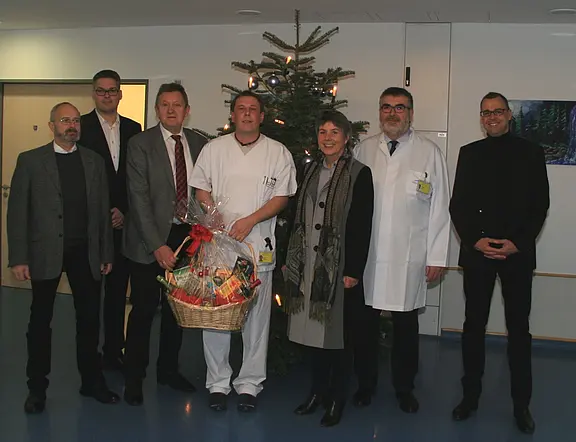 Gruppenfoto in einem Krankenhaus: Mehrere Mitarbeitende stehen vor einem Weihnachtsbaum. In der Mitte hält eine Pflegekraft einen großen Geschenkkorb mit roter Schleife. Daneben stehen Personen in Anzügen und zwei in weißen Kitteln. Festliche Stimmung, Übergabe eines Präsents.