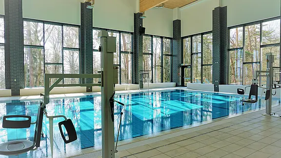 Helles Hallenbad mit großen Panoramafenstern. Ein rechteckiges Becken mit ruhigem, blauem Wasser. Mehrere Poollifte und Handläufe für barrierefreien Zugang. Draußen sind Bäume sichtbar.