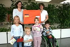 Eine Gruppe von fünf Personen, darunter drei Kinder, steht vor einer Pflanze. Eine Frau hält einen großen orangefarbenen Umschlag mit einem deutschen Aufkleber. Die Kinder halten kleine Karten und lächeln.