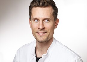 Dr. med. Jonathan Zühlke Porträt eines lächelnden Mannes in weißem Kittel, der direkt in die Kamera blickt, vor neutralem Hintergrund; wirkt wie medizinisches Personal.