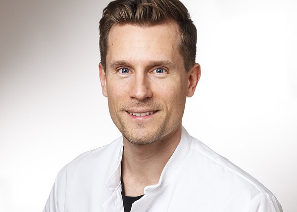 Porträt eines lächelnden Mannes in weißem Kittel, der direkt in die Kamera blickt, vor neutralem Hintergrund; wirkt wie medizinisches Personal.