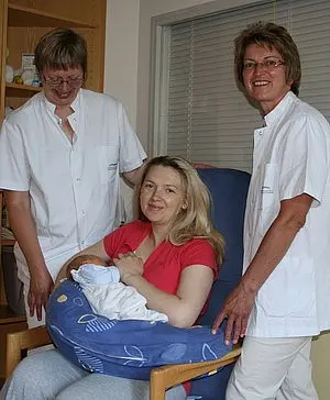 Die Abbildung zeigt eine Frau, die ein Neugeborenes in einem Stillkissen hält. Zwei Krankenpflegerinnen stehen neben ihr, lächeln und tragen weiße Oberbekleidung. Der Hintergrund ist hell und zeigt eine gemütliche, klinische Umgebung.