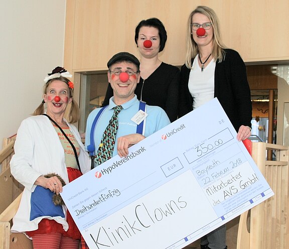 Vier Personen mit roten Clownsnasen präsentieren einen übergroßen Spendenscheck. Zwei sind als Klinikclowns verkleidet, zwei in Alltagskleidung. Auf dem Scheck steht „KlinikClowns“ und der Betrag 350 Euro. Fröhliche Übergabe in einem Innenraum.
