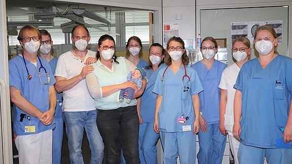 Gruppenfoto in einem Krankenhaus: Mehrere Pflegekräfte und Ärztinnen/Ärzte in blauen Kitteln und Masken. In der Mitte halten zwei Erwachsene ein Baby. Die Gruppe blickt zur Kamera; freundliche, feierliche Stimmung.