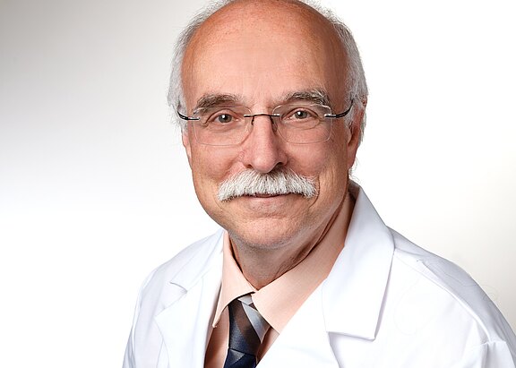 Porträt eines älteren Mannes mit Glatze, grauem Schnurrbart und Brille. Er trägt einen weißen Kittel über Hemd und Krawatte und lächelt freundlich in die Kamera vor neutralem Hintergrund.