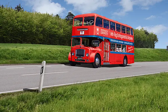 Roter Doppeldeckerbus im Retro-Stil fährt auf einer Landstraße. Große Aufschrift bewirbt einen Info-Tag zu Schlaganfall und Diabetes mit Präventionsbotschaften. Gesundheitskampagne unterwegs.