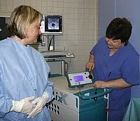 Eine medizinische Fachkraft in blauer Arbeitskleidung bedient ein Gerät, während eine andere Fachkraft in Schutzkleidung aufmerksam zuschaut. Beide scheinen in einem klinischen Umfeld zu arbeiten.