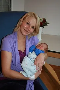 Eine lächelnde Frau sitzt auf einem Sessel und hält ein Neugeborenes in ihren Armen. Die Frau trägt ein lila Oberteil und hat blondes Haar. Das Baby ist in eine helle Decke gehüllt und schläft friedlich. Der Hintergrund zeigt einen schlichten Raum mit einer Blume in einer Vase.