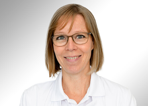 Porträt einer lächelnden Frau mit Brille und schulterlangem blondem Haar, in weißem Kittel, vor neutralem hellgrauen Hintergrund.