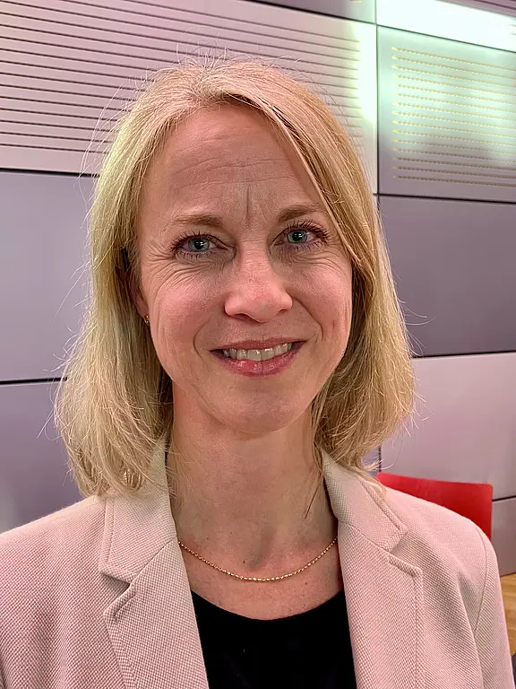 Sabine Kerner Porträtfoto einer lächelnden, blonden Frau mit schulterlangem Haar, hellem Blazer und schwarzem Oberteil; sie blickt in die Kamera vor neutralem Innenraumhintergrund.