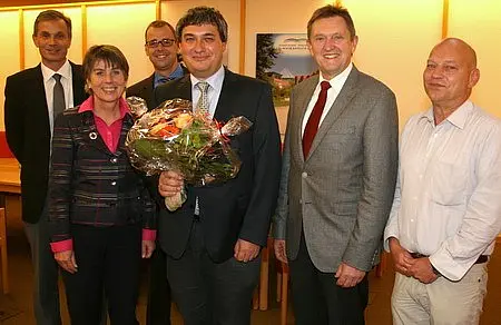Eine Gruppe von sechs Personen steht in einem Raum. Der Mann in der Mitte hält einen Blumenstrauß. Alle lächeln und scheinen an einem feierlichen Anlass teilzunehmen.