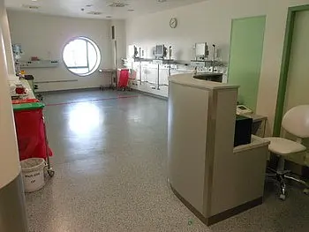 Eine helle, moderne Krankenhausumgebung mit einem Empfangstresen im Vordergrund. An der Wand befinden sich mehrere medizinische Geräte. Ein großes rundes Fenster lässt Tageslicht herein. Die Böden sind hell und der Raum wirkt aufgeräumt und sauber.
