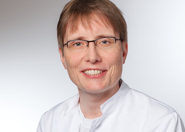 Ein lächelnder Arzt oder eine Ärztin mit kurzen, braunen Haaren trägt eine weiße medizinische Jacke und Brille. Der Hintergrund ist hell und neutral. Die Person vermittelt einen freundlichen und professionellen Eindruck.