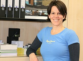 Petra Brückner Eine lächelnde Frau in einem blauen T-Shirt steht an einem Schreibtisch, der mit Ordnern und anderen Büromaterialien ausgestattet ist. Im Hintergrund sind Bilder von Autos und Landschaften zu sehen. Die Szene vermittelt eine freundliche und einladende Büroumgebung.