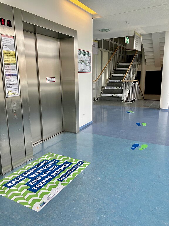 Flur mit Aufzug und Treppenhaus; Bodenplakat und Fußspuren weisen darauf hin, statt des Aufzugs die Treppe zu nutzen: „Nach oben ohne warten? Einfach die Treppen nehmen!“