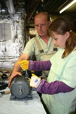 Ein Erwachsener und ein Kind arbeiten gemeinsam an einem Elektromotor. Der Erwachsene zeigt dem Kind, wie man mit Werkzeugen umgeht. Beide tragen Schutzhandschuhe und konzentrieren sich auf ihre Aufgabe in einer Werkstatt.