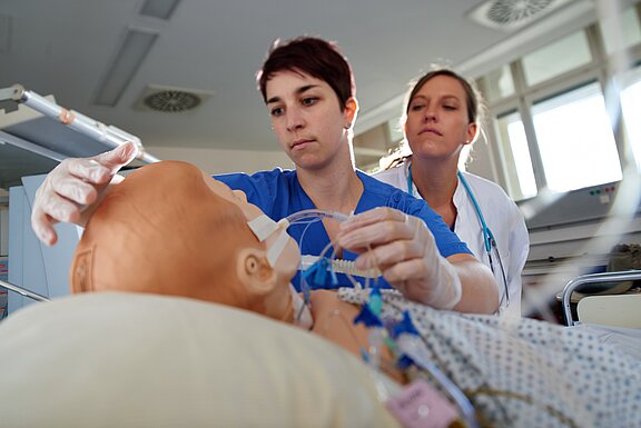 Zwei Fachkräfte in Krankenhausuniformen kümmern sich um eine Trainingspuppe. Eine Person platziert einen Schlauch an der Puppe, während die andere aufmerksam zuschaut. Die Szene zeigt eine medizinische Ausbildung oder Simulation zur Patientenversorgung.