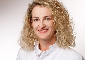 Aiste Matuleviciute-Stojanoska, Dr. med. Porträt einer freundlich lächelnden Frau mit lockigem blondem Haar in weißem Arztkittel vor hellem Hintergrund. Kopf und Oberkörper, direkter Blick in die Kamera.