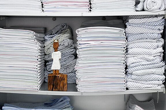 Mehrere Regale mit ordentlich gestapelter, weißer Bettwäsche und Handtüchern; in der Mitte steht eine kleine Holzfigur in weißem Gewand zwischen den Textilstapeln.