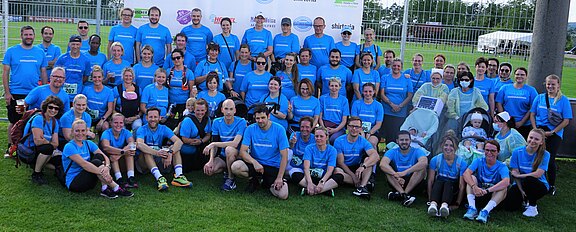 Große Gruppe Erwachsener in einheitlichen blauen Laufshirts posiert als Teamfoto auf einer Wiese. Einige sitzen oder knien, andere stehen; fröhliche Stimmung nach einem Sportlauf.