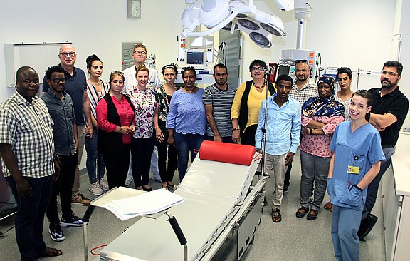 Gruppe von etwa zwanzig Erwachsenen posiert in einem hellen Krankenhaus-Behandlungsraum um eine Liege. Eine Pflegekraft in blauer OP-Kleidung lächelt vorn. Eindruck von Schulung, Team- oder Besuchergruppe in medizinischer Umgebung.