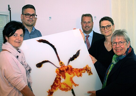 Fünf Personen posieren in einem Raum und halten gemeinsam ein großes Bild mit einer orange-braunen Orchidee in Nahaufnahme. Sie lächeln in die Kamera.