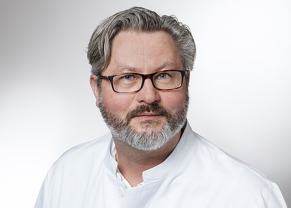 Porträt eines Mannes mit Brille und grauem Haar, kurzer Vollbart. Er trägt einen weißen Kittel und blickt ruhig in die Kamera vor neutralem Hintergrund.