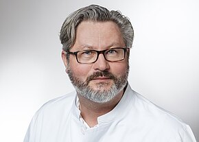 Porträt eines Mannes mit Brille und grauem Haar, kurzer Vollbart. Er trägt einen weißen Kittel und blickt ruhig in die Kamera vor neutralem Hintergrund.