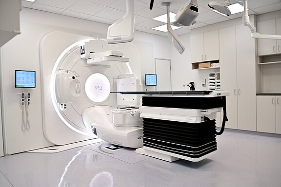 Heller Klinikraum mit großem, kreisförmigem Bestrahlungsgerät (Linearbeschleuniger) und verstellbarer Patientenliege; seitliche Monitore zur Steuerung. Moderne Radiotherapieanlage, einsatzbereit.