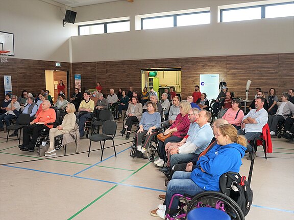 Große Gruppe Erwachsener sitzt in einer Turnhalle in Reihen und verfolgt aufmerksam eine Präsentation oder Rede; die Atmosphäre wirkt konzentriert und ruhig.