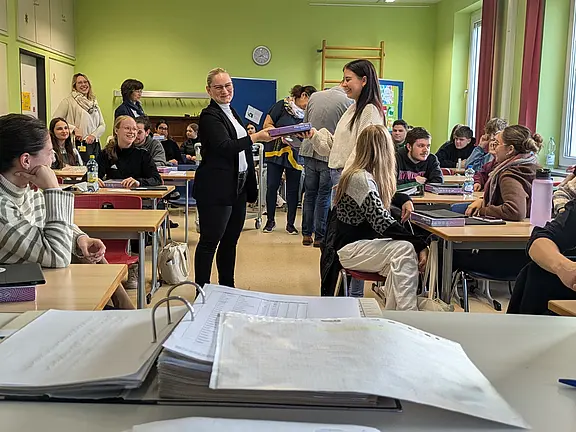 Klassenzimmer mit vielen Jugendlichen, sitzend an Tischen, einige lächeln. Im Mittelpunkt überreicht eine Lehrkraft einer Schülerin ein Buch; andere beobachten. Vordergrund unscharfe Unterlagen. Helle grüne Wände, Tageslicht durchs Fenster. Szene wirkt wie Verteilung von Unterrichtsmaterialien oder eine kleine Auszeichnung.