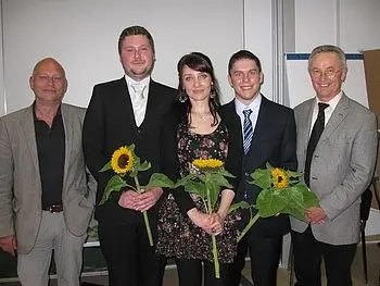 Fünf Personen stehen nebeneinander, lächeln und halten Sonnenblumen in den Händen. Sie tragen formelle Kleidung. Der Hintergrund ist neutral gehalten. Die Aufnahme vermittelt ein Gefühl von Feierlichkeit und Zusammengehörigkeit.