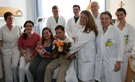 Eine Mutter hält ein neugeborenes Baby in den Armen, während ihr Partner mit einem Blumenstrauß lächelt. Um sie herum stehen mehrere medizinische Fachkräfte in weißen Kitteln, die ebenfalls freundlich schauen. Die Szene findet in einem Krankenhauszimmer statt.