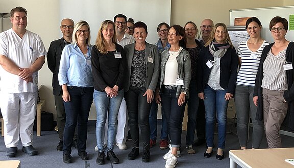 Eine Gruppe von 13 Personen steht in einem Raum. Sie lächeln und tragen unterschiedliche Kleidung, darunter Arbeitskleidung und legere Outfits. Im Hintergrund sind Möbel und eine Wandtafel sichtbar, die keine wichtigen Informationen enthält. Die Szene vermittelt eine freundliche und professionelle Atmosphäre.