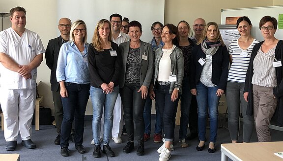 Eine Gruppe von 13 Personen steht in einem Raum. Sie lächeln und tragen unterschiedliche Kleidung, darunter Arbeitskleidung und legere Outfits. Im Hintergrund sind Möbel und eine Wandtafel sichtbar, die keine wichtigen Informationen enthält. Die Szene vermittelt eine freundliche und professionelle Atmosphäre.