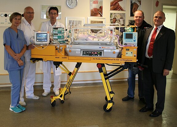 Im Krankenhaus steht eine gelbe mobile Transportinkubator-Einheit für Frühgeborene und Neugeborene, ausgestattet mit Monitoren und Beatmung. Mehrere Fachkräfte stehen daneben und demonstrieren das Gerät.