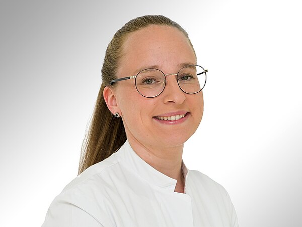 Ein lächelnde Person mit Brille trägt eine weiße Arztkittel. Der Hintergrund ist hell und weich verschwommen, was den Fokus auf das Gesicht der Person lenkt.