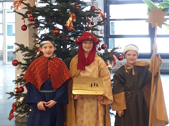 Drei Kinder in Königsgewändern posieren vor einem geschmückten Weihnachtsbaum. Sie stellen die Heiligen Drei Könige dar; eines hält einen Sternstab, ein anderes eine goldene Tafel mit Krippensilhouette. Die Szene vermittelt weihnachtliche Sternsinger-Tradition.