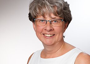 Sandra Braun Porträt einer lächelnden Frau mit kurzen, grauen Haaren, die eine brille trägt. Sie hat einen freundlichen Ausdruck und trägt ein ärmelloses, helles Oberteil. Der Hintergrund ist neutral und hell.
