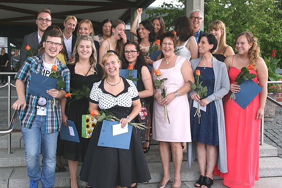 Gruppe junger Erwachsener in festlicher Kleidung posiert lachend draußen nach einer Abschlussfeier. Mehrere halten Urkunden und einzelne Rosen in der Hand. Jubelnde Stimmung, Menschen stehen dicht zusammen auf Treppenstufen.