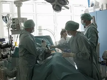Ein chirurgisches Team in einem Operationssaal führt einen Eingriff durch. Die Mitglieder tragen OP-Kleidung und Masken. Ein Patient liegt auf dem Operationstisch, umgeben von medizinischem Equipment und Monitoren.