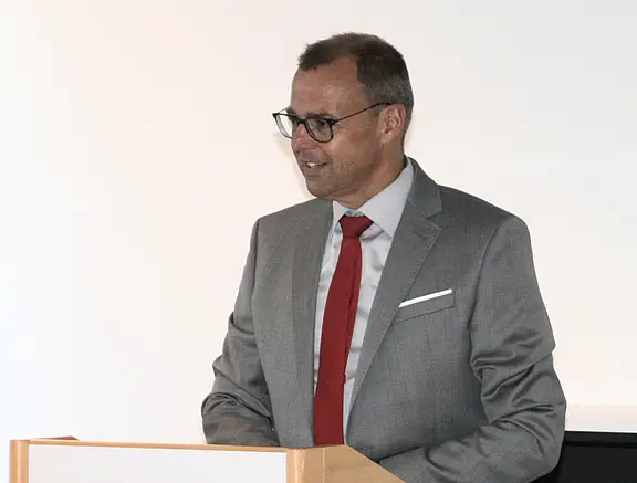 Ein Mann im grauen Anzug mit roter Krawatte spricht am Rednerpult. Er trägt eine Brille; neutraler, heller Hintergrund.