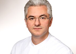 Filip Chkripeski Ein Mann mit grauen Haaren und Brille trägt ein weißes Hemd. Er schaut direkt in die Kamera. Der Hintergrund ist hell und einfarbig.