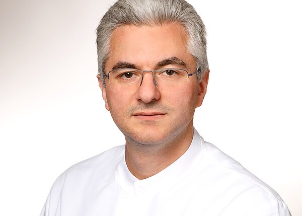 Ein Mann mit grauen Haaren und Brille trägt ein weißes Hemd. Er schaut direkt in die Kamera. Der Hintergrund ist hell und einfarbig.