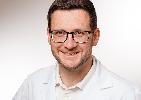 Ein junger Mann mit Brille lächelt freundlich in die Kamera. Er trägt einen weißen Kittel und ein helles Shirt. Der Hintergrund ist neutral gehalten und unterstützt den professionellen Eindruck des Bildes.