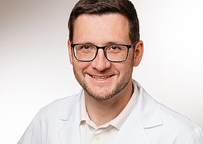 Ein junger Mann mit Brille lächelt freundlich in die Kamera. Er trägt einen weißen Kittel und ein helles Shirt. Der Hintergrund ist neutral gehalten und unterstützt den professionellen Eindruck des Bildes.
