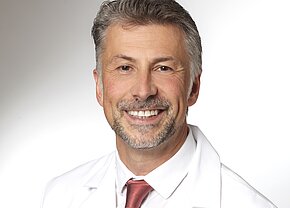 Univ.-Prof. Dr. med. Jürgen Dolderer Ein lächelnder Arzt in weißem Kittel mit einem roten Hemd darunter. Sein Haarschnitt ist kurz, und er hat einen gepflegten Bart. Der Hintergrund ist hell und schlicht, was den Fokus auf sein freundliches Gesicht lenkt.